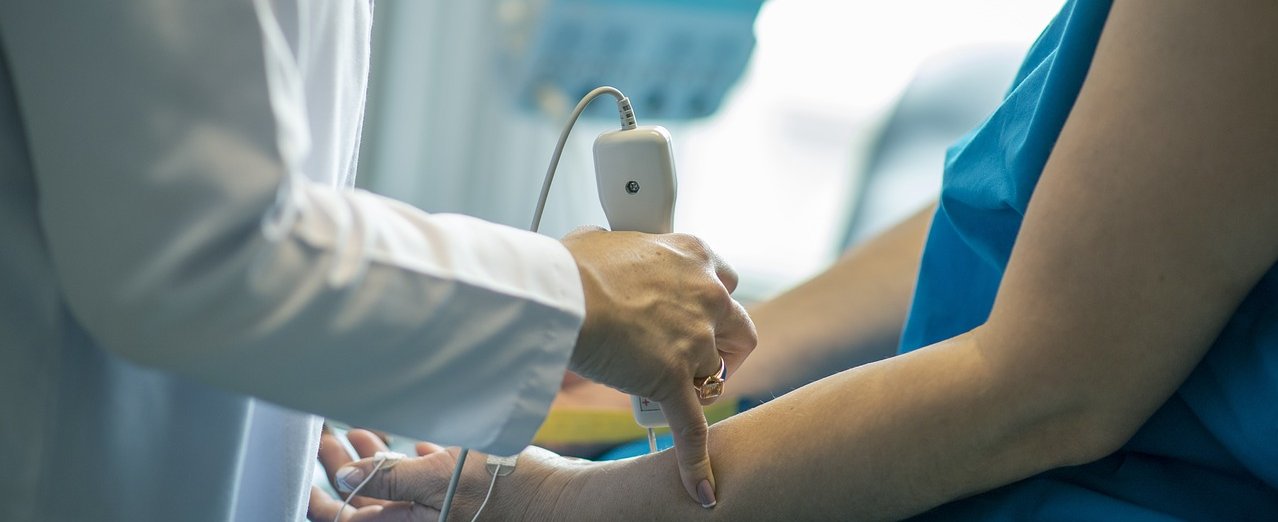 Eine Person in weißem Kittel steht vor einem Patienten und führt ein Gerät über den Arm. 